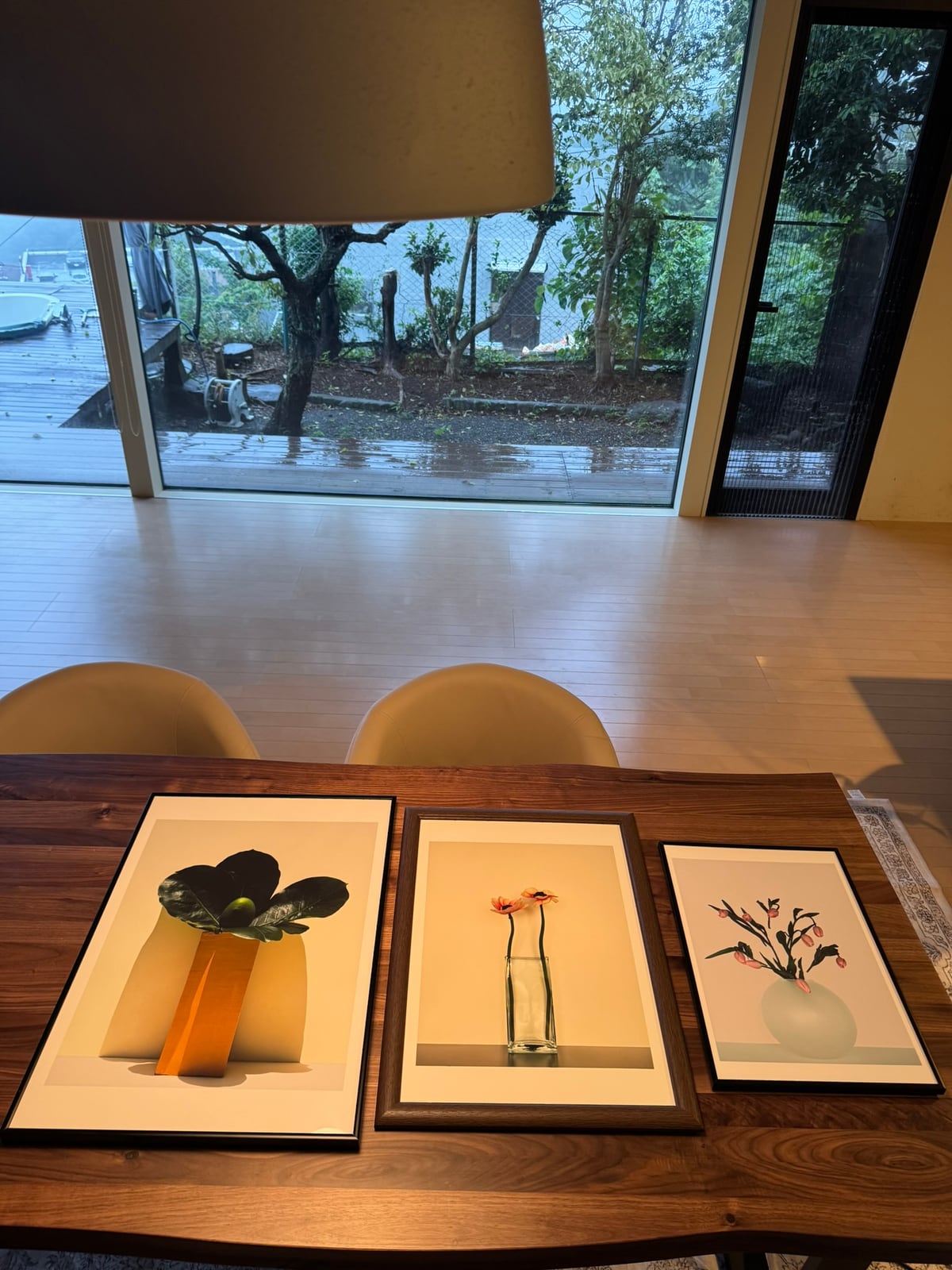 Three framed prints on a walnut table in front of a rain-washed garden window